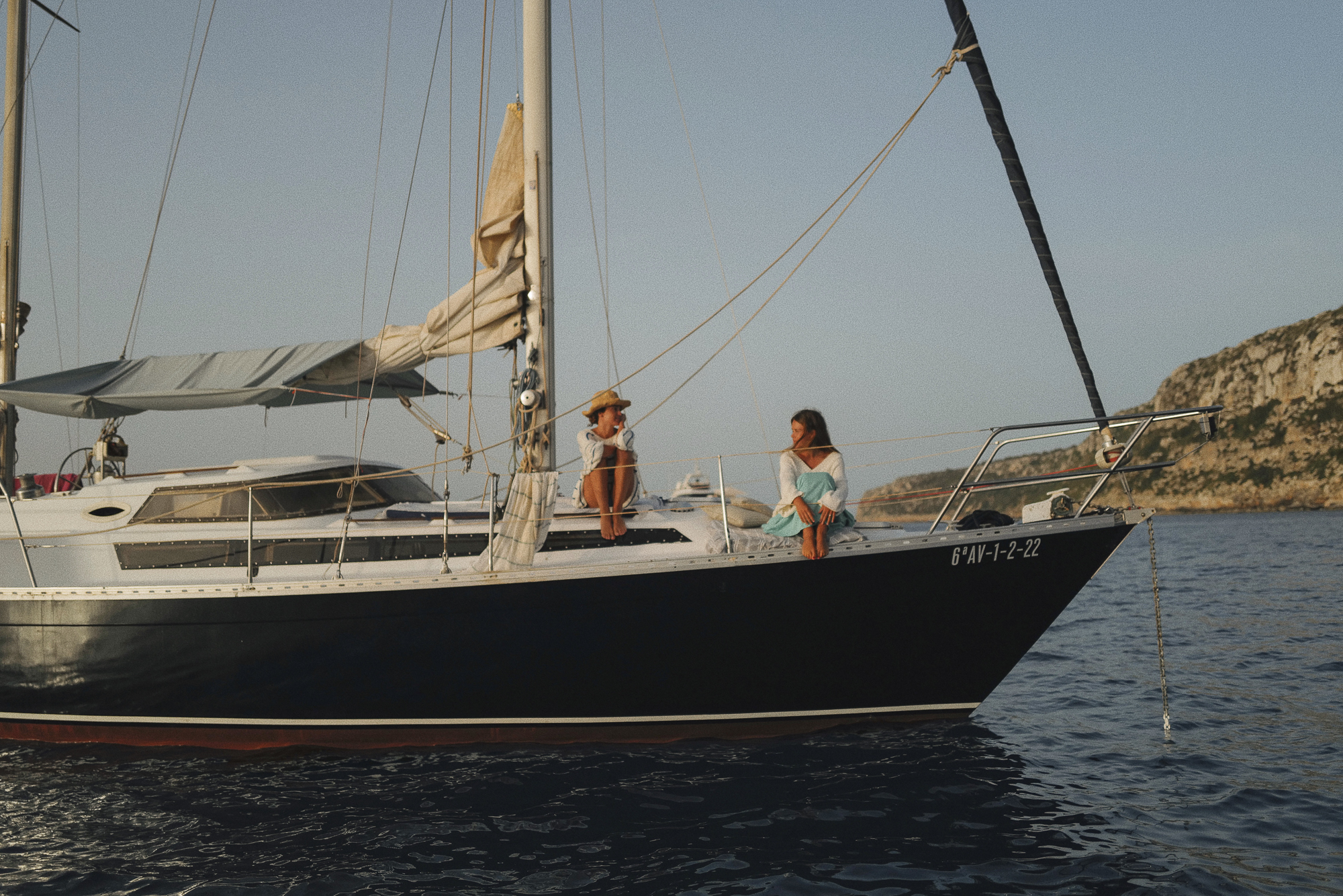 Two friends engaging in conversation on the deck of the 'CALIU' with the scenic coastline of Formentera in the background, embodying the vessel’s intimate and friendly sailing experience. Full day sailing tour in es Caló