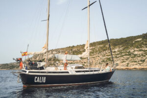 Passengers relaxing on the deck of the 'CALIU', moored off the rugged coast of Formentera, epitomizing the sailboat's promise of a safe and idyllic seafaring adventure.