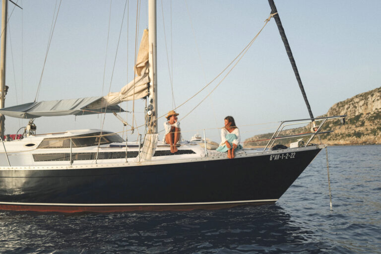 The Caliu is anchored in Es Caló, Formentera. Side view of the sailboat. Two girls are sitting, talking and enjoying the moment. Full day sailing tour in Formentera