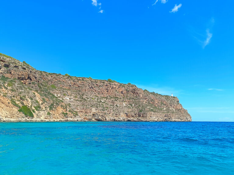 Calm turquoise waters of Cala Codolar lead the eye to the majestic La Mola lighthouse perched atop the cliff, a beacon during our coastal explorations.